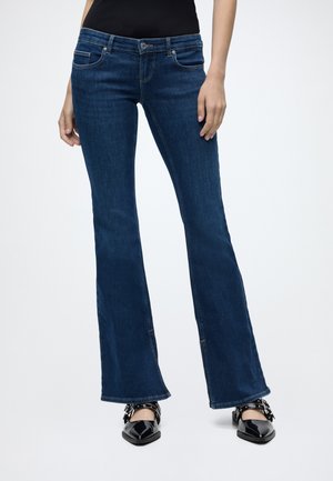 Femme portant un jean flare bleu foncé avec des ballerines noires à bouts pointus, debout sur un fond blanc.