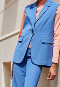 Blå skräddarsydd väst med slag, enknappsstängning och två framfickor, lager på en orange ribbad topp, tillsammans med blå byxor.