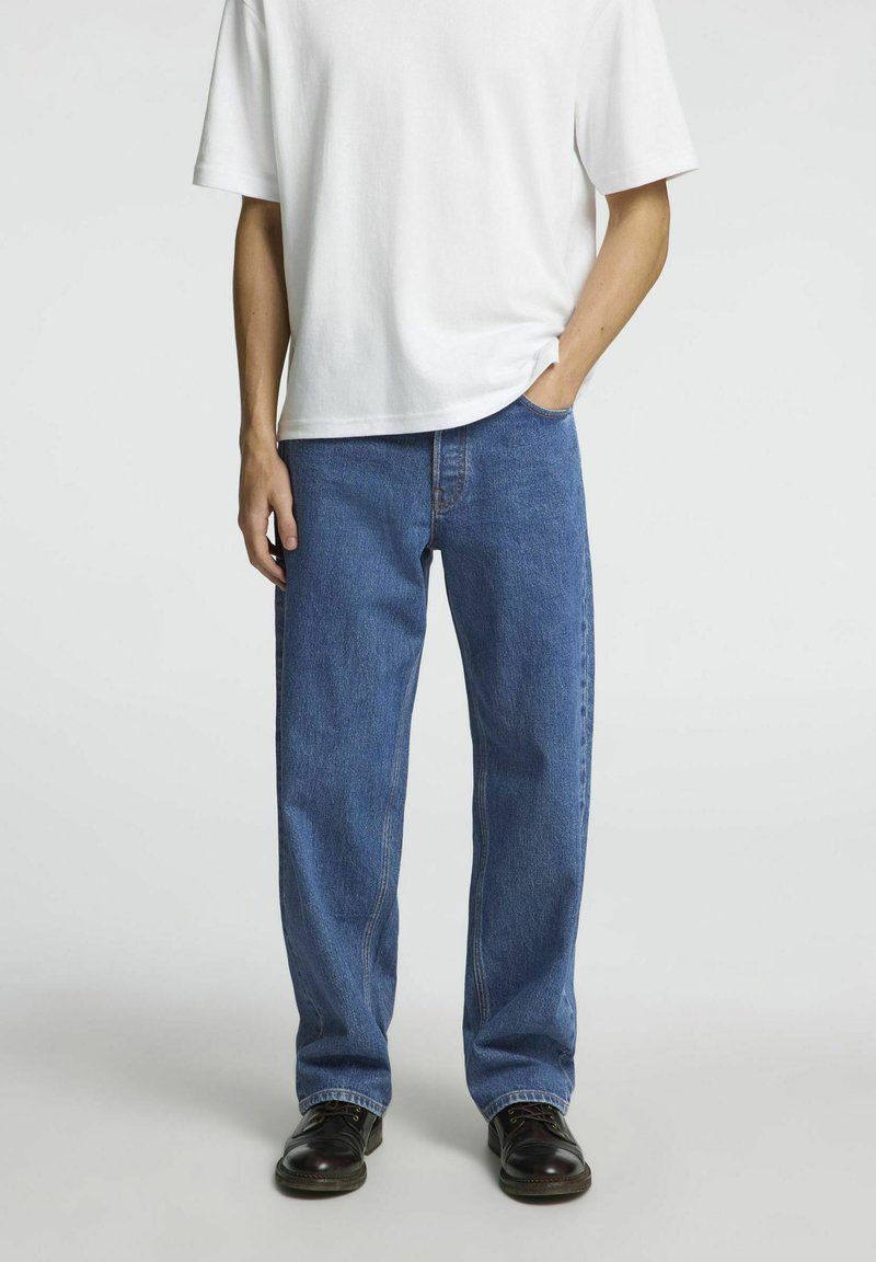 Blå denimjeans med en avslappnad passform och rak bendesign, ihop med en enkel vit t-shirt i bomull. Klädseln har enkla linjer och minimal detaljer.
