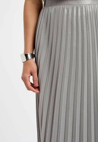 Abito grigio plissettato realizzato in tessuto lucido, con una cintura liscia e un braccialetto in argento metallico al polso sinistro.