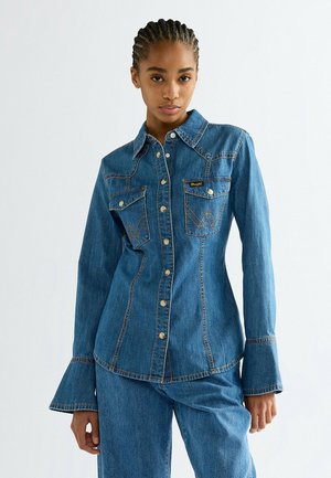 Frau mit geflochtenen Haaren, die ein tailliertes blaues Jeanshemd mit Druckknöpfen, zwei Brusttaschen, ausgestellten Manschetten und passende Jeanshosen trägt.