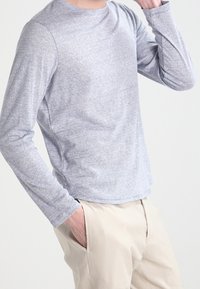 Chemise à manches longues et col rond de couleur gris clair, fabriquée en tissu doux avec une texture subtile. Elle présente un ourlet arrondi et des manches ajustées.