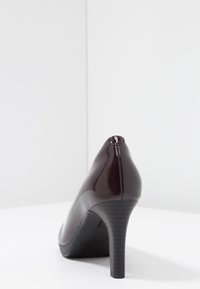 Pump lucido di un elegante bordeaux scuro con punta affusolata e tacco nero sottile. Superficie liscia con una cucitura sul retro.