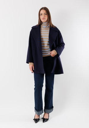 Cappotto di lana navy con ampi revers, dolcevita beige e blu a righe, jeans in denim scuro con risvolti e décolleté in vitello lucido nero con fiocchi.