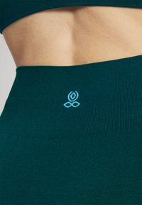 Leggings de sport teal avec un tissu lisse et extensible; présente un logo bleu clair à la taille. Pas de motifs ni d'accents supplémentaires.