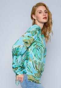 Leichtes, türkisfarbenes Blusenoberteil mit einem Blattmuster. Verfügt über lange Ärmel, einen lockeren Schnitt und weichen, durchsichtigen Stoff. Akzente in Grün und Braun.