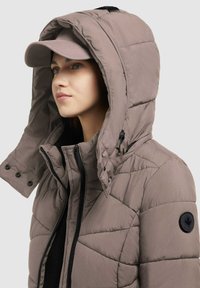 Jachetă puff în taupe deschis cu design matlasat cusut. Dispune de o glugă mare, nasturi negri și o pata pe mânecă stângă. Este completată de o șapcă bej.