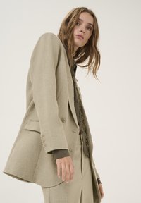 Lys beige skræddersyet blazer med en struktureret overflade, to knapper og sidelommer, parret med en mørk olivengrøn skjorte nedenunder.