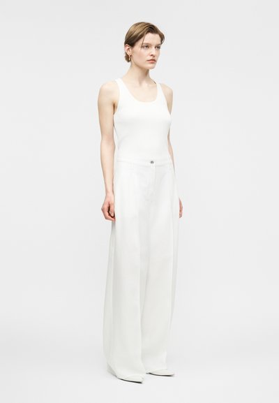 Γυναίκα που στέκεται φορώντας λευκό αμάνικο τοπ, λευκό παντελόνι φαρδιάς γραμμής και λευκά μυτερά παπούτσια, μπροστά από απλό ανοιχτό φόντο.