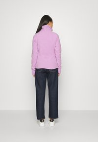 Lila Fleecejacke mit hohem Kragen, kombiniert mit dunkelblauen weiten Jeans und weißen Sneakern mit schwarzen Akzenten, von hinten gesehen.