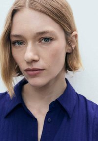 Camicetta a maniche corte di colore blu navy con tessuto texture, dotata di colletto e dettagli con bottoni, indossata da una persona con capelli lunghi fino alle spalle.