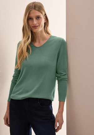 Groene, lichte trui met een V-hals en lange mouwen, met ribgebreide manchetten en een losse pasvorm, gecombineerd met donkerblauwe jeans.