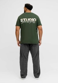Camiseta de algodón verde con un gran texto gráfico blanco "STUDIO Jack & Jones" en la espalda, combinada con pantalones de pana grises.