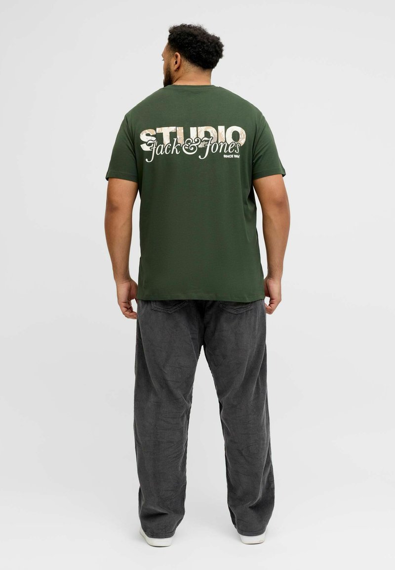 Camiseta de algodón verde con un gran texto gráfico blanco "STUDIO Jack & Jones" en la espalda, combinada con pantalones de pana grises.