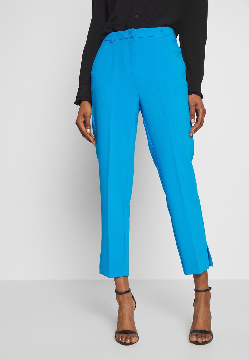 Kobieta w jasnoniebieskich, skrojonych w krótkim stylu spodniach, czarnym długim topie i czarnych szpilkach z nadgarstkiem, stojąca na jednolitym tle.