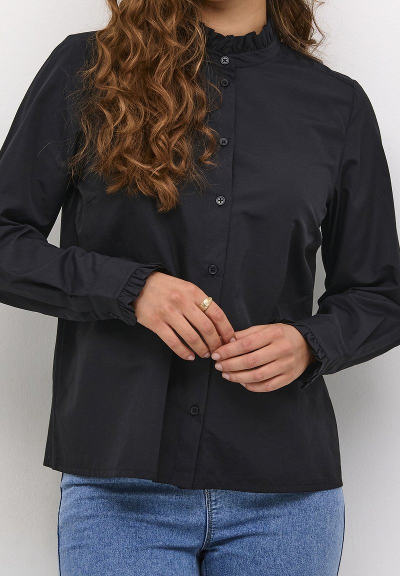Femme aux cheveux bouclés portant un chemisier noir boutonné avec un col à volants et des poignets, un jean bleu et une bague en or à son doigt.