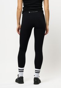 Leggings neri con una texture liscia, dotati di un logo sul bordo in vita, abbinati a sneaker nere e calzini bianchi con strisce nere.
