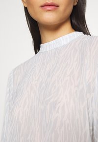 Femme portant un chemisier léger à col montant avec des motifs abstraits subtils ressemblant à des zèbres, ne montrant que le cou et le bas du visage.