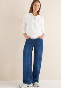 Sweat-shirt blanc à capuche avec cordons lacés à l'avant, associé à un jean en denim bleu foncé à coupe ample. Le mannequin porte des baskets gris clair.