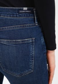 Mörkblå denimjeans med en standard bakficka och en synlig varumärkesetikett. Tyget visar en lätt textur och blekna detaljer.