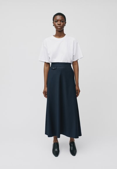 Maglietta bianca a maniche corte abbinata a una gonna maxi blu navy a vita alta con dettaglio di bottoni laterali; il modello indossa mule nere.