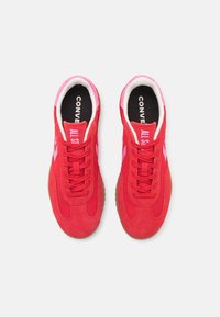 Baskets en suède rouge avec des accents roses, présentant un design à lacets, des semelles intérieures noires avec du texte blanc, et une semelle extérieure en gomme texturée.