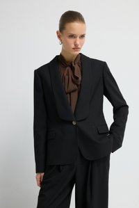 Zwart blazer met krijtstreepjes, een sjaalkraag, enkele knoopsluiting en twee voorzakken; gecombineerd met een bruine blouse met strik.