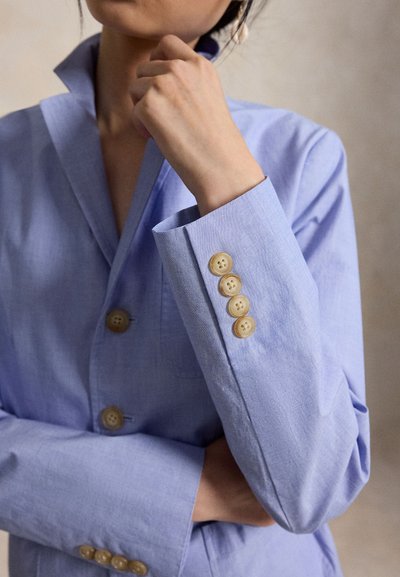 Γυναίκα με ανοιχτό μπλε σακάκι με μπεζ κουμπιά, το χέρι κοντά στο σαγόνι, ποζάροντας μπροστά σε ουδέτερο φόντο.