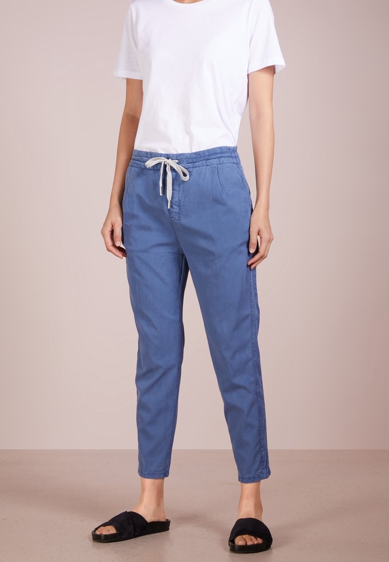 Personne portant un pantalon bleu avec cordon, une chemise blanche à manches courtes et des sandales noires slip-on, debout sur un sol beige.