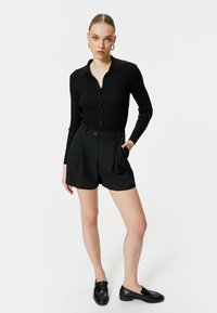 Schwarze gerippte langärmlige Strickjacke mit Knöpfen, kombiniert mit schwarzen hochgeschnittenen Shorts, beide aus strukturiertem Material. Schwarze Schuhe runden das Outfit ab.