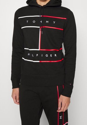 Man wearing black Tommy Hilfiger hoodie and matching black pants with red and white stripes and brand name details.