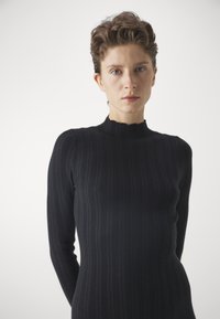 Pulover negru tricotat cu un guler înalt, ondulat și mâneci lungi. Materialul are o textură netedă și o siluetă ajustată.