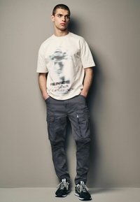 Cremefarvet grafisk t-shirt med et sløret statuedesign. Grå cargo bukser med flere lommer. Sorte sneakers med hvide snørebånd.