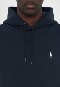 Hanorac de culoare bleumarin, cu un buzunar tip cangur, șnururi ajustabile și un mic logo alb pe pieptul stâng. Material dintr-un amestec de bumbac texturat.