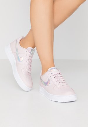 Nogi skrzyżowane, ubrane w jasnoróżowe sneakersy Nike z opalizującym logo i białą podeszwą, stojące na jasnoszarej podłodze.