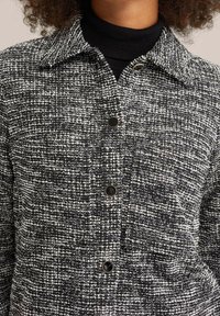 Svart och vit mönstrad texturerad tygjacka med krage och knappstängning, med fyra runda svarta knappar framtill.