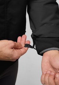 Zwarte jas met een gestructureerde stof en fleece manchetten, met een knoopsluiting en verstelbare band op de mouw. Hand houdt de band vast.
