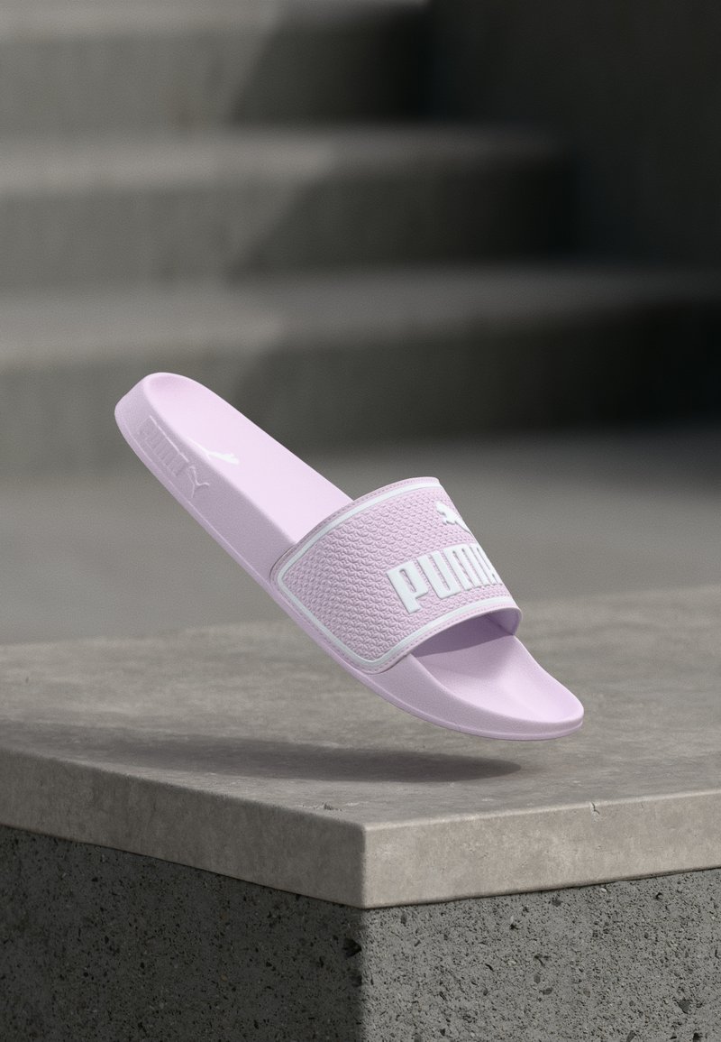 Sandale slide Puma couleur violet clair flottant au-dessus d'un rebord en béton avec des marches en béton floues en arrière-plan.