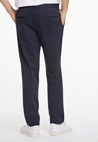 Pantalones azul marino de tela suave con un corte entallado, que cuentan con dos bolsillos traseros con cierre de botón y una línea a lo largo de la pierna. Zapatillas deportivas blancas.