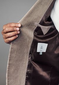 Beige Cordjacke mit glattem Satinfutter. Innentasche und Pflegeetikett sichtbar. Hand, die an den Revers greift.