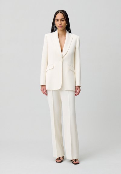 Traje blanco a medida que consiste en un blazer largo y pantalones de pierna ancha, con un escote en V profundo y dos bolsillos frontales. Textura suave.