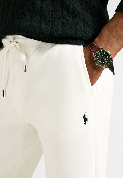 Λευκή φόρμα με κορδόνι στη μέση και μαύρο κεντημένο λογότυπο αλόγου. Συνδυάζεται με πράσινο πλεκτό πουλόβερ με ανάγλυφα σχέδια και ρολόι με πράσινο καντράν.