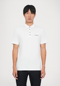 Hvid polo t-shirt med knapkrave, korte ærmer og et sort logo på brystet. Enkel design, bomuldsstof, glat tekstur.