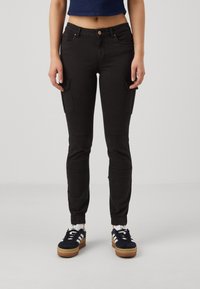 Svarta cargojoggers med elastiska muddar, framfickor och extra sidfickor, tillverkade av ett strukturerat tyg. Matchas med svarta sneakers.