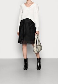 Weißer Strickpullover mit V-Ausschnitt, schwarzer plissierter Midi-Rock, schwarze Ankle Boots und eine Handtasche mit Schlangenhautmuster.