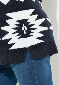 Maglione a maglia bianco e nero con motivo geometrico, fessura laterale all'orlo, abbinato a jeans denim blu. La texture sembra morbida.