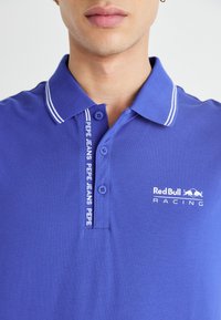 Modrá polokošeľa s bielymi pruhmi na golieri, so začiatkom na dvoch gombíkoch a logami Red Bull Racing a Pepe Jeans na hrudi.