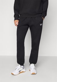 Calças de estilo sweat em algodão preto com punhos elásticos, apresentando um pequeno logótipo branco da Reebok do lado esquerdo. Usadas com sapatilhas brancas.
