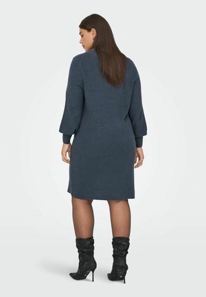 Robe pull en maille de couleur bleu foncé, avec de longues manches ballons, une coupe décontractée et une longueur mi-thigh. Associée à des bottes noires à talons.
