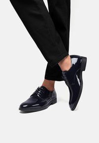 Scarpe in pelle verniciata blu navy con finitura liscia, chiusura con lacci chiusi e tacco basso, abbinate a pantaloni neri.